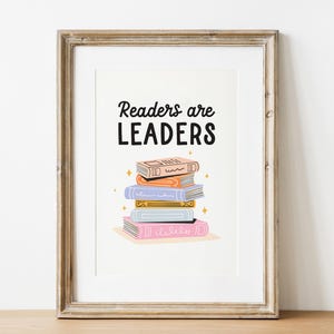 Könnte beinhalten: Gerahmter Kunstdruck mit dem Text "Readers are LEADERS" über einem Stapel bunter Bücher. Die Bücher sind in Orange-, Blau-, Gelb- und Rosatönen gehalten, mit goldenen Sternakzenten. Der Rahmen ist hellbraun.