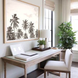 May include: A sepia-toned framed print of palm trees hangs above a wooden desk. A white lamp, small plant, and a tray with decor sit on the desk. A white chair is positioned in front of the desk, creating a serene office space.