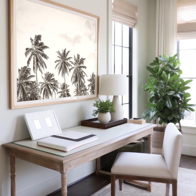 May include: A sepia-toned framed print of palm trees hangs above a wooden desk. A white lamp, small plant, and a tray with decor sit on the desk. A white chair is positioned in front of the desk, creating a serene office space.