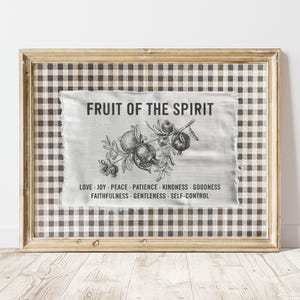 May include: Framed artwork featuring a gray and white gingham pattern. The artwork includes a linen-like fabric with the text "FRUIT OF THE SPIRIT" and a botanical illustration. Below the text are words like "LOVE" and "JOY".