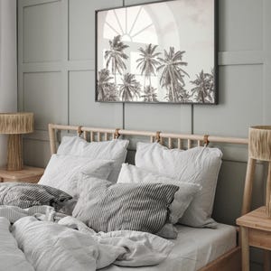 May include: A bedroom scene featuring a bed with white linens and striped pillows. Above the bed hangs a framed black and white photograph of palm trees. Two wooden lamps with woven shades flank the bed.