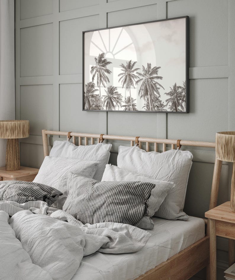 May include: A bedroom scene featuring a bed with white linens and striped pillows. Above the bed hangs a framed black and white photograph of palm trees. Two wooden lamps with woven shades flank the bed.