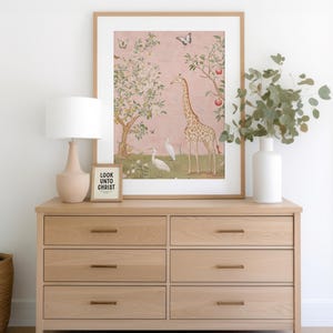 May include: A light oak dresser with six drawers, brass handles, and a framed print of a giraffe and birds on a pink background. A small framed sign reads "LOOK UNTO CHRIST". A beige lamp and white vase with greenery complete the scene.