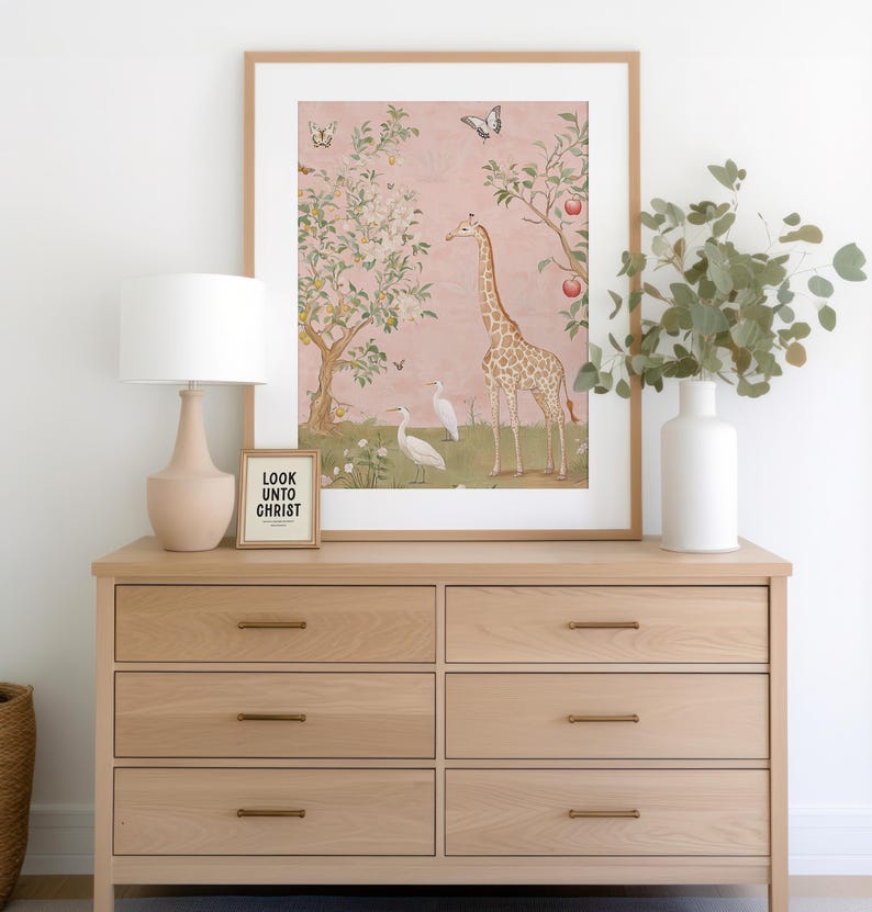 May include: A light oak dresser with six drawers, brass handles, and a framed print of a giraffe and birds on a pink background. A small framed sign reads "LOOK UNTO CHRIST". A beige lamp and white vase with greenery complete the scene.
