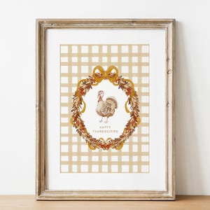 May include: Framed Thanksgiving art print. The artwork features a turkey inside a wreath of autumn leaves and ribbons, set against a beige plaid background. The words "Happy Thanksgiving" are printed below the turkey. The frame is a light brown color.