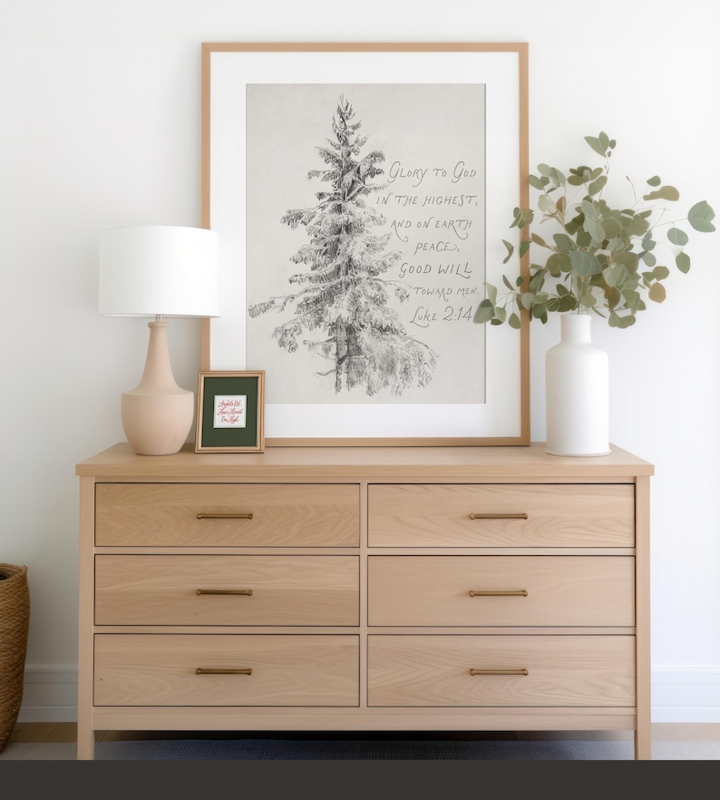May include: A light oak dresser with six drawers and brass handles, featuring a framed print of a detailed tree sketch. The print includes the text "Glory to God in the Highest, and on Earth Peace, Good Will toward Men, Luke 2:14." A lamp and vase are also on the dresser.