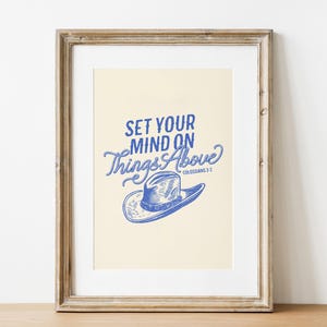 May include: Framed art print with a blue cowboy hat illustration and the text "SET YOUR MIND ON Things Above" in a vintage style. The print is in a wooden frame and is displayed on a light-colored background.