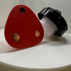 May include: A red leather triangle with three gold metal circles and a heart design. The triangle is sitting on a white surface next to a black and white bottle cap.