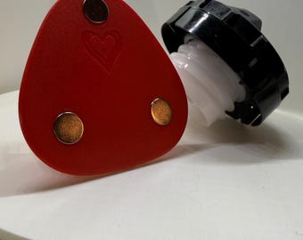 May include: A red leather triangle with three gold metal circles and a heart design. The triangle is sitting on a white surface next to a black and white bottle cap.
