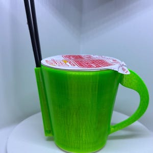 Könnte beinhalten: Eine grüne, im 3D-Druck hergestellte Tasse mit einem weißen Deckel und Stäbchen, die aus der Oberseite herausragen. Die Tasse steht auf einem weißen Drehteller.