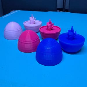 May include: Five 3D printed eggs, each with a different color and a small 3D printed hand on top. The eggs are purple, pink, blue, and two shades of blue.