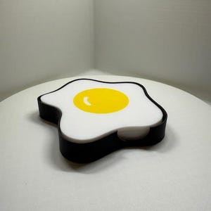 Puede incluir: Un posavasos en forma de huevo blanco y negro con una yema amarilla. El posavasos está hecho de un material suave y flexible y tiene un borde elevado.