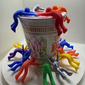 以下が含まれることがあります： カラフルな3Dプリントされたフィギュアのグループが、白いラーメンカップの側面にしがみついています。カップはカラフルな絵で飾られており、「日清」と「ラーメン」の文字が見えます。フィギュアは様々なポーズをとっており、中には手を伸ばしているもの、ぶら下がっているもの、カップの側面にしがみついているものがあります。