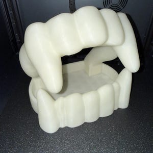 May include: A 3D-printed, off-white set of vampire teeth. The top and bottom rows of teeth are connected, forming a hinged box. The teeth are large and detailed, with visible ridges and curves. The object is sitting on a dark, textured surface.