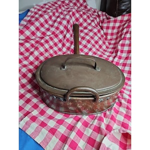 May include: An antique copper oval casserole dish with a lid and a long handle. The dish has a handle on the lid and on the side. The copper has a warm, aged patina. The dish is sitting on a red and white checkered cloth.