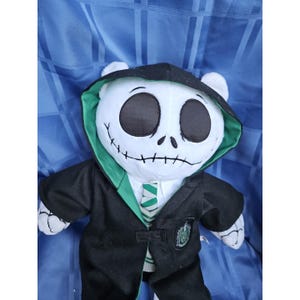 May include: A plush toy resembling Jack Skellington from The Nightmare Before Christmas, wearing a black robe with green trim and a Hogwarts crest. The bear has a stitched mouth and large black eyes, with white fur on its head and paws.