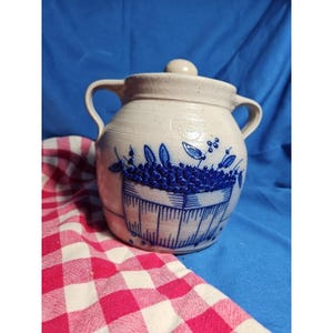 Puede incluir: Un tarro de gres con tapa, con un diseño azul y blanco de arándanos en una cesta. El tarro tiene dos asas y un pomo en la tapa. El fondo es una superficie azul con un paño a cuadros rojos y blancos.