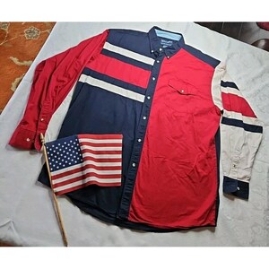 Pode incluir: Uma camisa de botão de manga comprida com um esquema de cores patriótico. A camisa apresenta painéis e listras vermelhas, brancas e azul marinho. Uma bandeira americana é colocada perto da camisa. A camisa tem um bolso na frente.