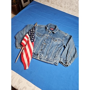 May include: A denim jacket with a button-up front and pockets, paired with a small American flag. The jacket is a medium wash blue color. The flag has red and white stripes and a blue field with white stars.