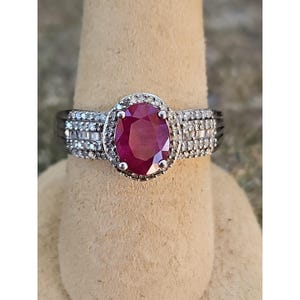 May include: A silver ring featuring a large, oval-cut ruby gemstone. The ruby is surrounded by a halo of smaller, clear gemstones. The ring's band is also adorned with additional clear gemstones, creating a sparkling effect.