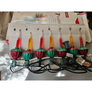 Könnte beinhalten: Ein Set aus sieben NOMA-Blasenlichtern mit abwechselnd roten und gelben flüssigkeitsgefüllten Glasröhren auf gerippten roten und grünen Kunststoffsockeln. Die Lichter sind durch ein schwarzes Stromkabel verbunden und auf einem Karton befestigt.