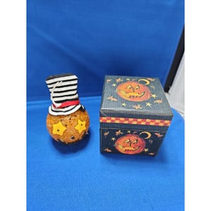 May include: A round orange textured pumpkin figure wearing a black and white striped top hat with a red band and yellow star-shaped eyes, positioned next to a square black box decorated with orange jack-o'-lantern faces and stars.