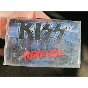 May include: A cassette tape featuring the band KISS. The album title "REVENGE" is in red, with the band's name in a silver, stylized font. The background is a blue and gray industrial design.
