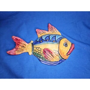 May include: A ceramic fish wall ornament featuring a yellow head and body with blue, green, and red painted details. The fish has a prominent red tail fin, a blue dorsal fin with dark blue accents, and a textured scale pattern on its side.