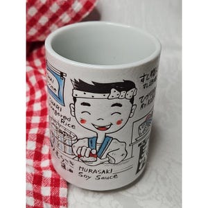 Puede incluir: Taza de cerámica blanca con una ilustración de un chef de sushi japonés. La taza presenta un chef sonriente con texto que incluye "Chari Vinegared Rice" y "Murasaki Soy Sauce". El diseño es en blanco y negro con detalles en rojo.