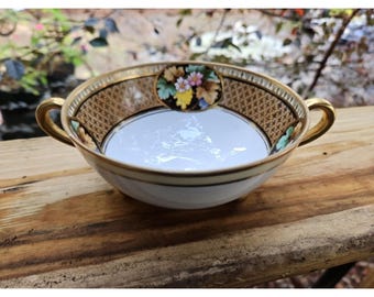 May include: A white and gold porcelain soup bowl with two gold handles. The bowl features a decorative brown and gold pattern around the rim and a floral design in the center. The bowl is on a wooden surface.