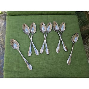 May include: A set of seven polished stainless steel spoons arranged on a surface. Three pairs of spoons are crossed in an X-shape, while one single spoon is positioned to the left and another single spoon is placed to the right.
