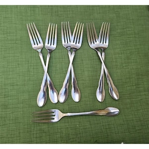 May include: A set of seven silver-toned metal forks. Three pairs of forks are arranged in a crossed pattern, while one single fork is positioned horizontally below them. Each fork features four tines and a smooth, rounded handle design.