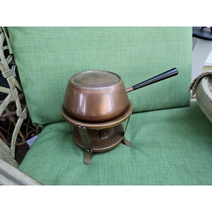 May include: A copper fondue pot with a black handle and a matching stand. The pot is round with a lid and sits on a metal stand with three legs. The pot is approximately 6 inches in diameter.