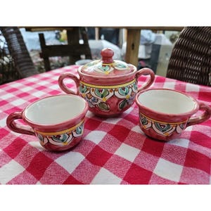 Op de afbeelding: Een keramische theeset met een suikerpot en twee kopjes. De set is beschilderd met een rode achtergrond en kleurrijke bloemmotieven. De kopjes en de suikerpot hebben oren en staan op een rood-wit geruit tafelkleed.