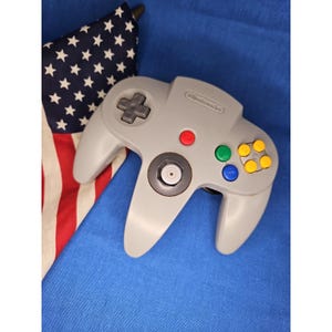 Puede incluir: Un mando de videoconsola Nintendo 64 de color gris con un stick analógico central, una cruceta, un botón de inicio rojo y cuatro botones de acción de colores verde, azul y amarillo. El logotipo de Nintendo está grabado en la parte superior.