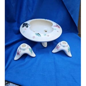 Puede incluir: Una jardinera de cerámica blanca con diseño de mariposas, con un cuenco redondo sostenido por tres patas. También hay dos candelabros a juego con motivos de mariposas. Las mariposas son en tonos rosa, azul y negro.