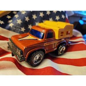 Könnte beinhalten: Ein Vintage-Spielzeug-LKW mit einer Wohnkabine. Der LKW ist hauptsächlich orange mit braunen und gelben Akzenten und einer gelben Kabine. Der Hintergrund ist eine amerikanische Flagge.