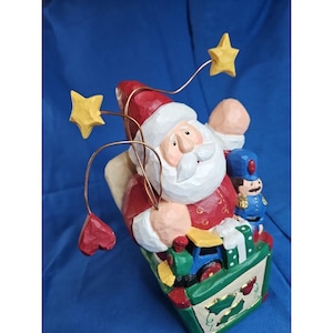May include: A carved Santa Claus figurine with a white beard and red suit, holding a sack filled with toys. The figurine has a whimsical design with yellow stars and a red heart attached to copper wire. A toy soldier is also visible.