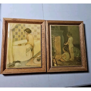 May include: Two rectangular wall plaques framed in light brown wood. The left plaque depicts a child in a bathroom with a tiled wall and bathtub. The right plaque shows two children standing and sitting near a fireplace with a metal screen.