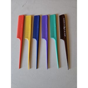 May include: An assortment of colorful hair combs in red, yellow, blue, purple, teal, and brown. The combs have long, pointed handles and fine teeth. The brown comb has text that reads "GOLDILOCKS 5 BY Krest DELRIN."