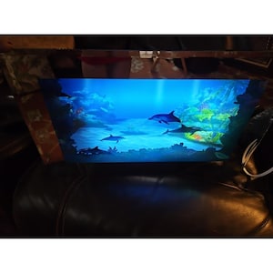 Può includere: Un'opera d'arte rettangolare incorniciata che raffigura una scena sottomarina con diversi delfini che nuotano in un oceano blu. L'opera presenta una tavolozza di colori vivaci con sfumature di blu, verde e bianco, creando un ambiente acquatico sereno e accattivante.