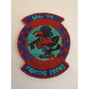Op de afbeelding: Geborduurde patch met een rode rand en een centraal ontwerp met een zwarte kraai die een wapen vasthoudt. De patch bevat de tekst "60th TFS" bovenaan en "Fighting Crows" onderaan, met blauwe en groene accenten.