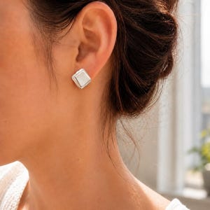 May include: A small, square-shaped silver stud earring with a smooth, polished finish. The earring features a simple, geometric design that sits flush against the earlobe, reflecting light on its flat surface and slightly beveled edges.