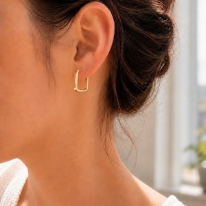 May include: A gold-toned hoop earring with a rectangular shape and rounded corners. The earring features a smooth, polished metallic finish and is secured through the earlobe with a thin post, creating a clean and simple geometric design.