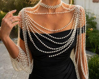 Könnte beinhalten: Ein schwarzes schulterfreies Kleid, verziert mit einer aufwendigen, mehrlagigen Perlen-Körperkette. Das Accessoire besteht aus einem Perlenhalsband im Choker-Stil mit zahlreichen drapierten weißen Perlensträngen über Schultern und Armen.