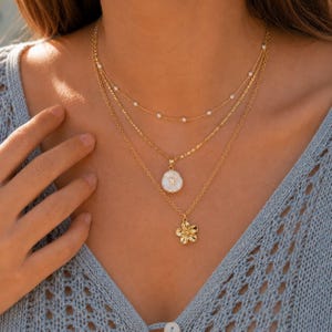 May include: A layered gold-tone necklace set consisting of three chains. The top chain features small white beads, the middle chain holds a round white shell-like pendant, and the bottom chain features a gold-tone flower-shaped charm.