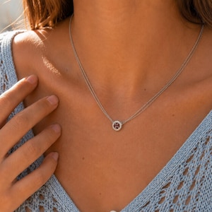 May include: A double-strand silver-toned necklace featuring a circular pendant. The pendant has a dark red centre stone surrounded by a halo of small, clear, sparkling stones. The necklace rests against a light blue, open-knit textured garment.