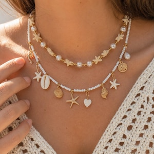 May include: A layered necklace set featuring a gold-tone chain with white beads, cream-coloured starfish, and round pearls. The second strand is a white beaded necklace with gold-tone charms including starfish, seashells, a heart, and a dolphin.
