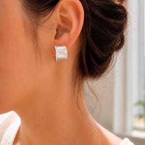 May include: A silver-toned earring with a smooth, curved, and wide band design. The earring features a polished metallic finish that reflects light, creating a subtle sheen across its concave surface. It is a simple, solid piece of jewellery.
