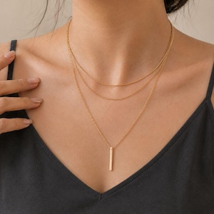 May include: A layered gold necklace set. The top layer is a delicate chain, the middle layer is a thin bar, and the bottom layer features a vertical rectangular pendant. The necklaces are worn over a black top.
